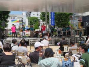 屋外のステージ上で演奏しているところを観客が座っている後方から撮影した写真