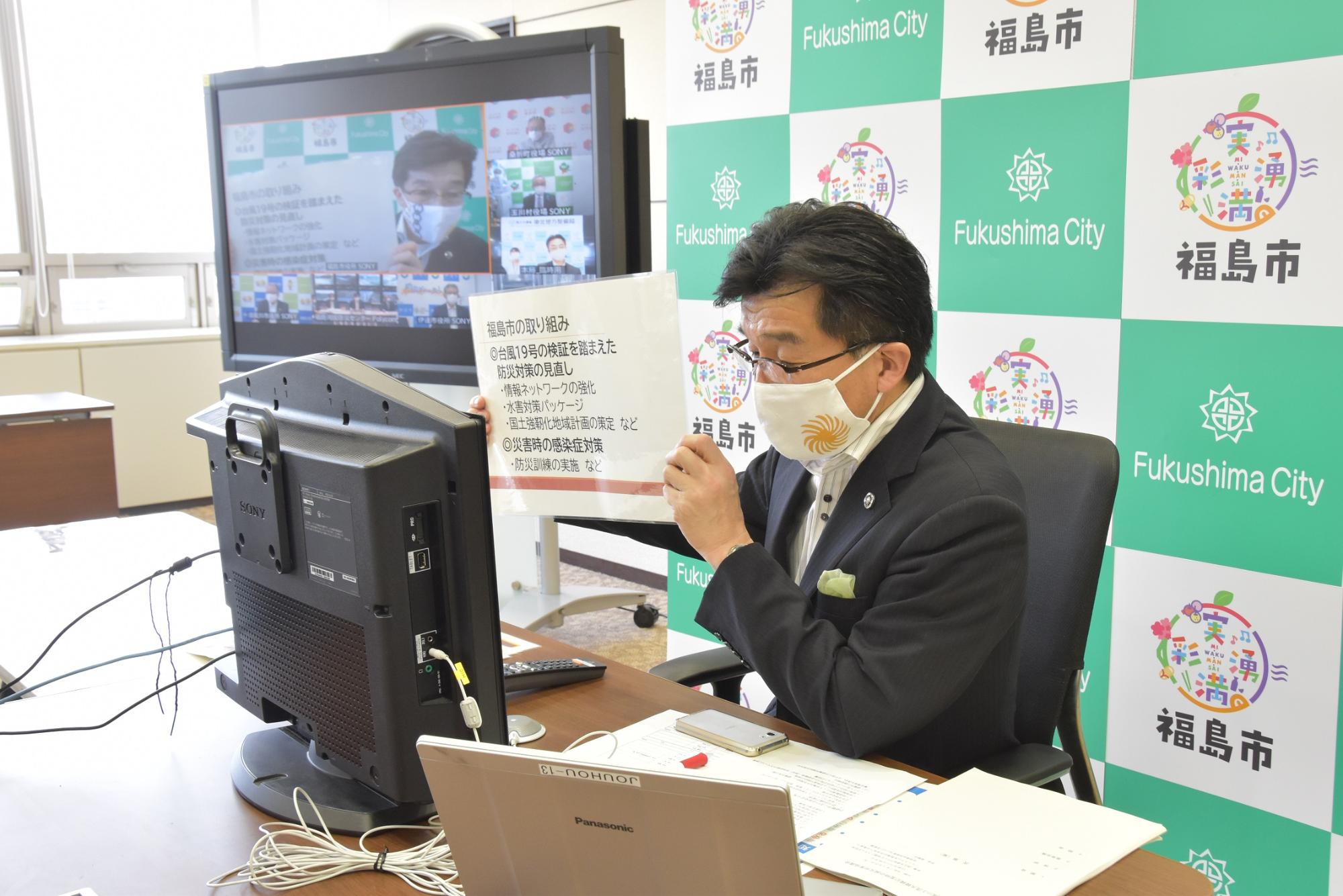 福島市のバックボードの前で市長が座って文字が書かれた紙を持ちWEB会議をしている様子が市長の横に設置されたモニターに映し出されている写真