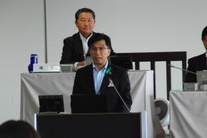 後方で男性が見ている中、市長が演台に立って話をしている写真