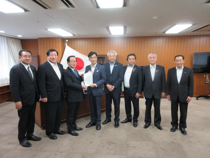 議員の執務室内で市長を含む男性8名が立ち、市長と男性で要望書を持っている写真