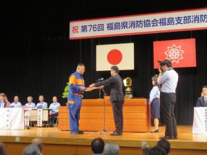 来賓の方々が座っているステージ上で市長が作業服を着た消防団員に賞状を渡している写真