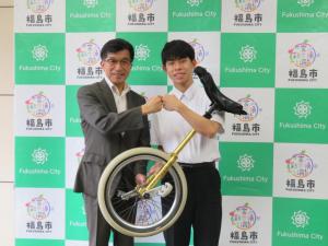 バックパネルの前で、市長と制服姿の男子生徒が一輪車を抱え、グータッチをしている写真