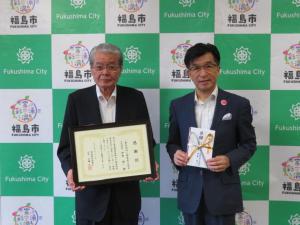 バックパネルの前で、市長が目録を持ち、男性が額縁に入った賞状を持って立っている写真