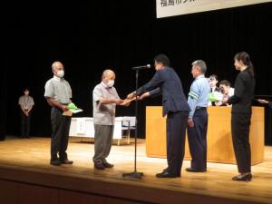 ステージ上で関係者の方が見ている中、市長が高齢の男性に委任状を手渡している写真