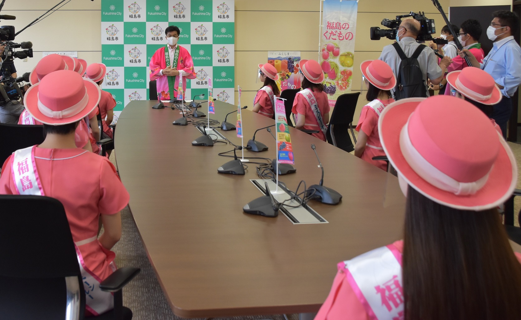 福島市のバックボードの前で市長が立ち、その前に設置された縦長の机を回りにピンク色のワンピースと帽子を着用した女性達が座り、市長の話を聞いている写真