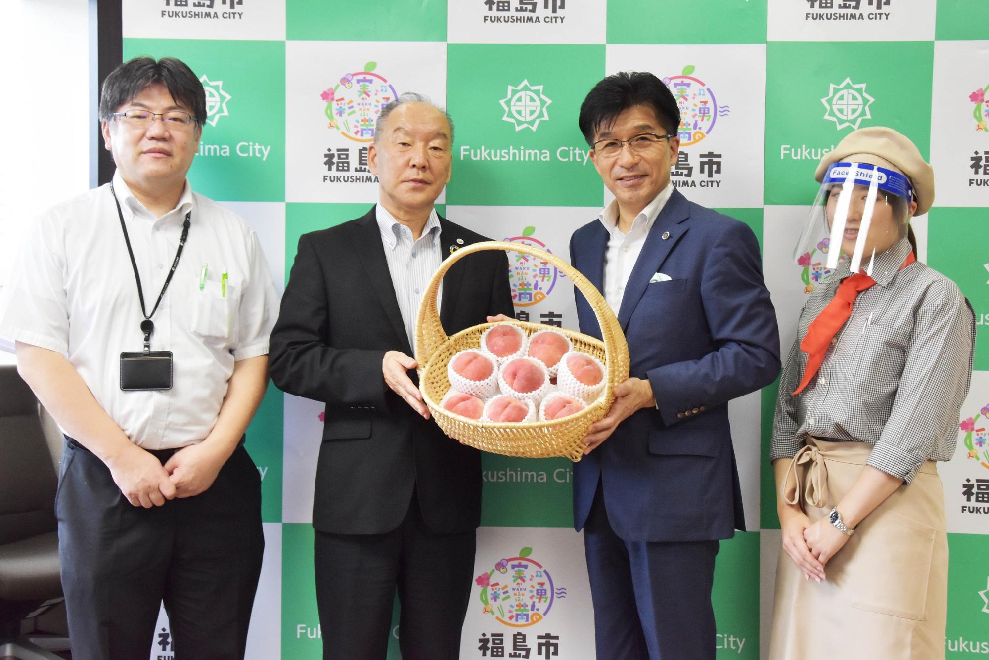 福島市のバックボードの前でフェイスシールドを付けた女性と市長と男性2名が横並びに立ち、男性と市長が桃の入った果物かごを一緒に持ち、記念撮影している写真