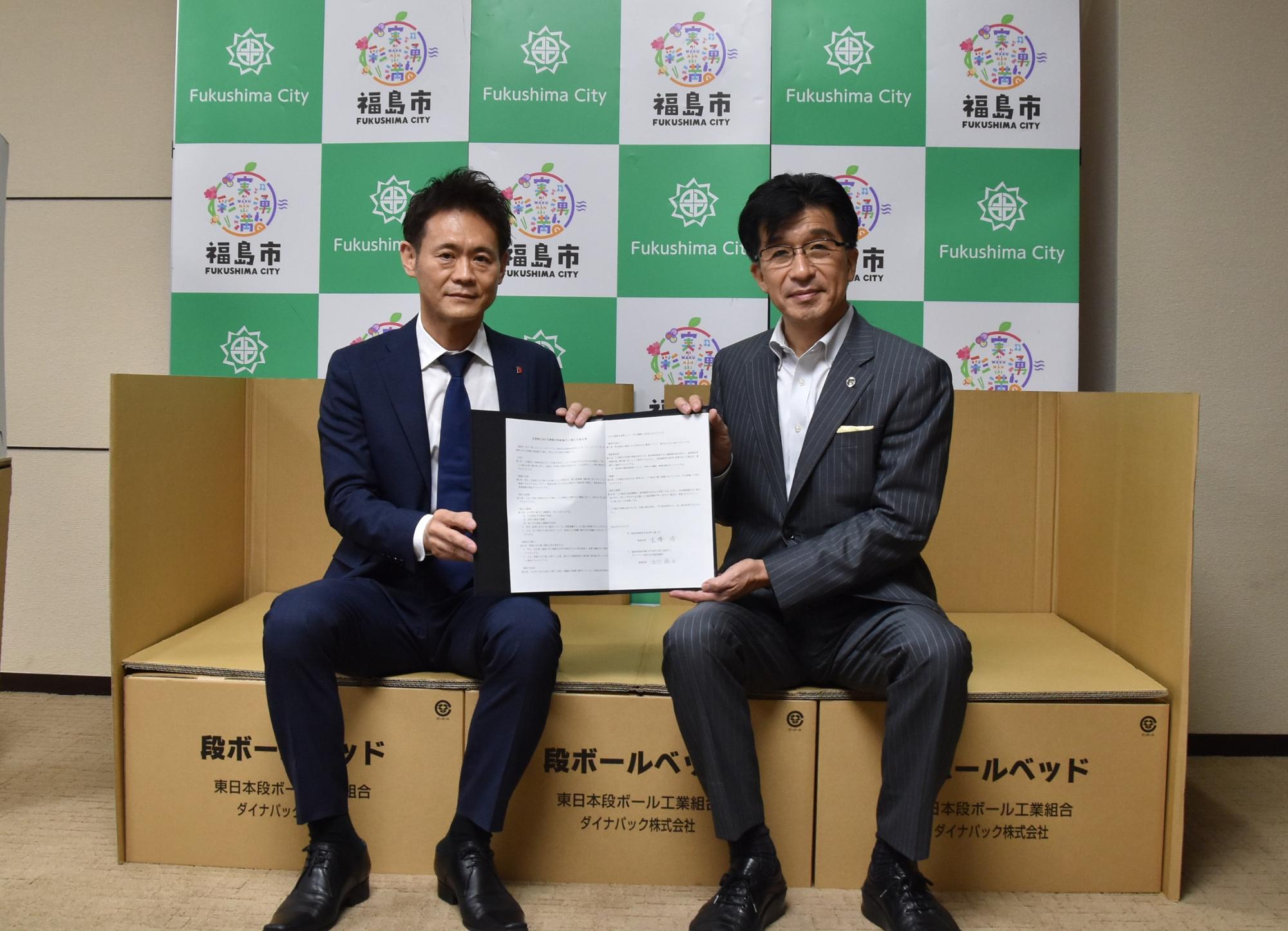 福島市のバックパネル前で、ダンボールベッドに腰掛け、協定書を持つ出口事業所長と市長の写真