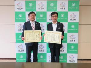 表彰状を持った2人の男性が福島市のバックボードの前で記念撮影をしている写真