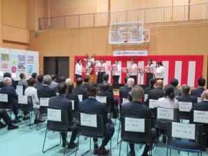 紅白幕が飾られた舞台で演奏をしている人達をパイプ椅子に座っている関係者の人達を後方から写した落成式の写真