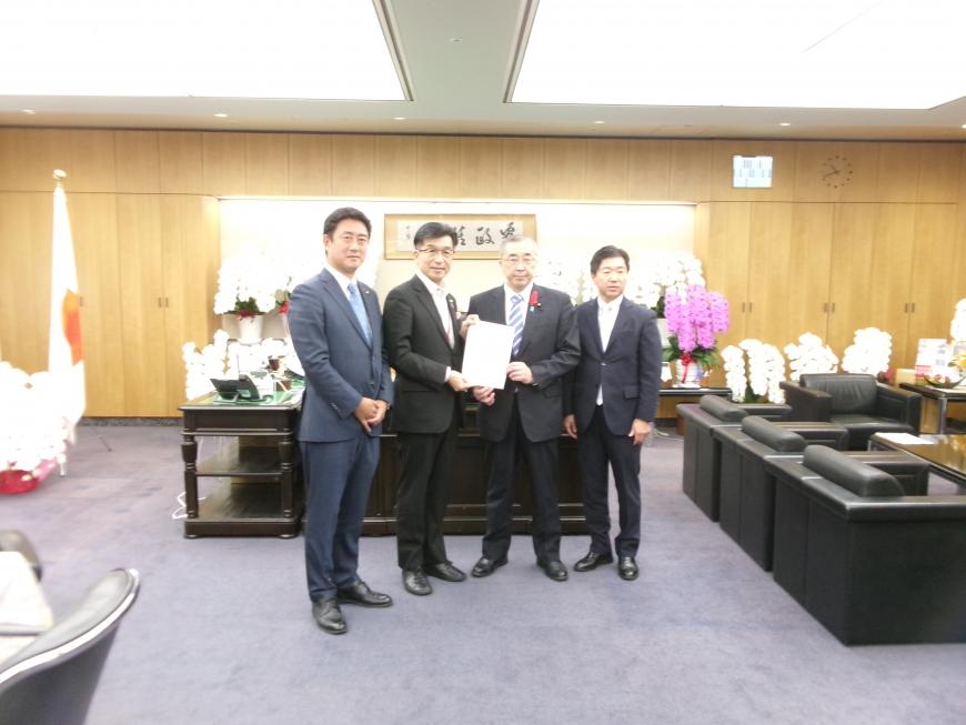 胡蝶蘭が飾られた執務室内で、市長を含む男性4名が横一列に並び、市長と男性で要望書を持っている写真