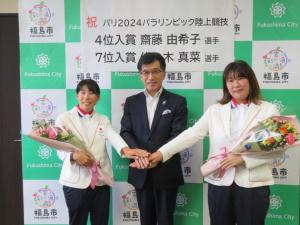 白のジャケット姿の女性2名が花束を持ち市長と手を重ねて記念撮影をしている写真