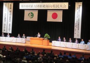 第34回暴力団根絶福島県民大会の幕が飾られた舞台の演台で挨拶をしている市長と左右に設置された机に並んで座っている関係者の人達を写した写真