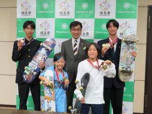 男子学生2名と小学生の女の子2名がスケートボードを持ち、首にはメダルをかけて市長と記念撮影をしている写真