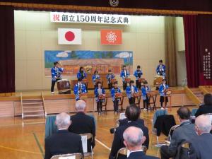 体育館の舞台で太鼓の演奏をしている生徒たちとそれを見ている人達の後方から写した写真