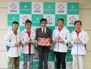 白のお揃いのジャンバーを着た男性4名と中央に箱入りのりんごをもった市長が1列に並んでいる記念撮影の写真