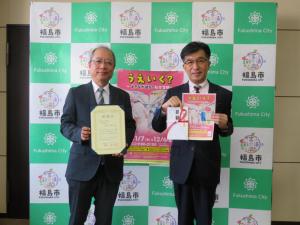 福島市のバックボードの前で男性が賞状を持ち市長が寄附金とチラシを持ちながら記念撮影をしている写真