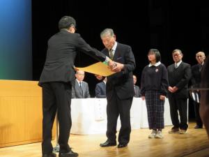 左側に表彰される人が1列に並び、先頭の男性に表彰状を手渡す市長の後方から写した写真