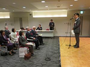 スタンドマイクの前に立つ市長と1段下に並んで座っている表彰者の人達を横から写した写真