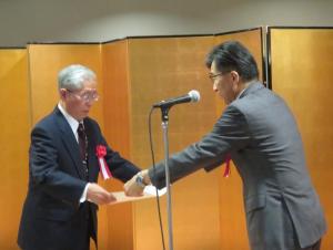 スタンドマイクの横で男性に賞状を手渡す市長の写真