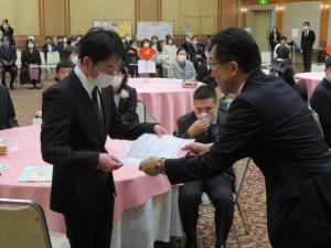 ピンク色のテーブルクロスが掛けられた丸テーブルの椅子から立ち上がったスーツ姿の男性が、市長から記念証書を手渡されている様子の写真