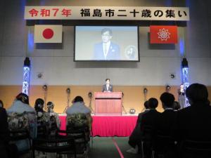 「令和7年 福島市二十歳の集い」と書かれた横断幕の下、国旗と市旗の間に市長が映し出されたスクリーンが設置されたステージの檀上で市長が挨拶を行っている写真