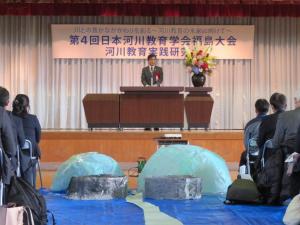 第4回日本河川教育学会福島大会の舞台の花が飾られた演台で挨拶をしている市長を正面から写した写真