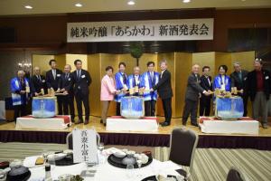 「純米吟醸「あらかわ」新酒発表会」と書かれた横断幕と金屛風が設置された小さなステージ上に3つの酒樽が置かれ、関係者たちが木槌を持ち並んでいる写真