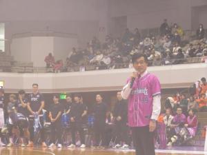 観客や選手たちが集まっている中、ピンク色のユニフォームを着た市長が右手でマイクを持ち挨拶を行っている様子の写真