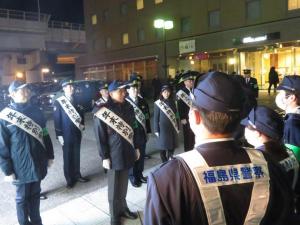 太陽が沈んだ夜の屋外で、紺色の制服を着用し肩からタスキをかけた警察官の方々が整列している写真