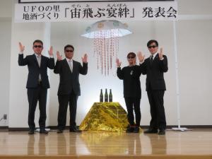 「UFOの里地酒づくり「宙飛ぶ宴絆(そらとぶ えんばん)」発表会」と書かれた横断幕の下にUFOの模型と金色の台の上に置かれた地酒の瓶が3本並び、両端にサングラスをかけた4名の関係者が両手を上げて並んでいる写真