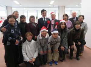 サンタの帽子を被った男性と胸章リボンをつけた市長を中央に他12人が並んでいる写真