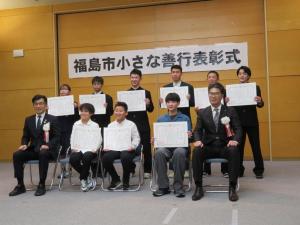 壁に「福島市小さな善行表彰式」と書かれた会場で、胸章リボンをつけた市長と関係者の男性、賞状を持った3人が前列の椅子に座り、後列に6人が立っている記念写真