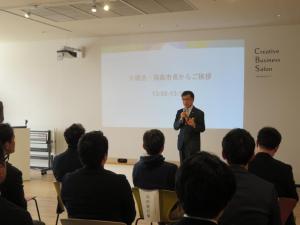 白い壁に資料の表紙が映し出された前に立つ市長が、向かい合って設置された椅子に座った参加者の人たちに向かって話をしている写真