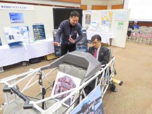 前後に座席が備わっている模型の後部座席に市長が座り、男性スタッフから話を聞いている様子の写真