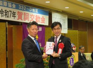 賀詞交歓会の会場で、ライオンズクラブの代表の男性と胸章リボンをつけた市長が一緒に目録を持ち並んでいる記念写真