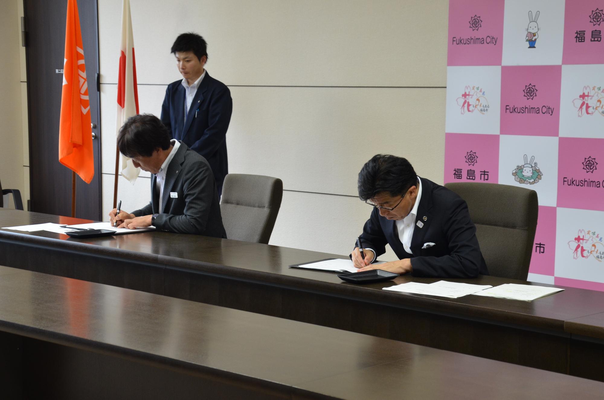 市長と関係者が椅子に座り、協定書に記入をしている写真