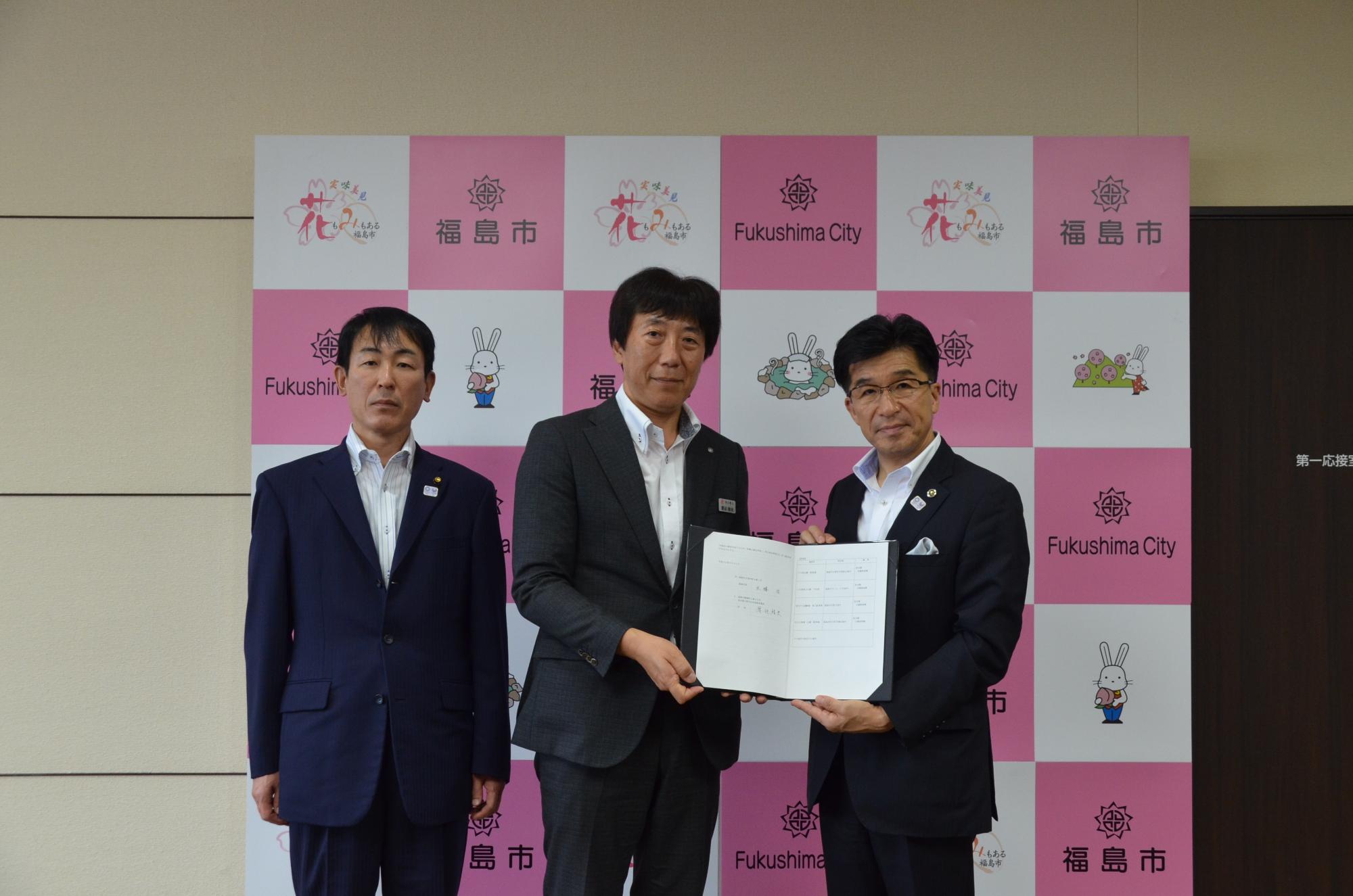 バックパネルの前で市長と男性が2名並び、市長と男性が協定書に手を添えて正面を向いている写真