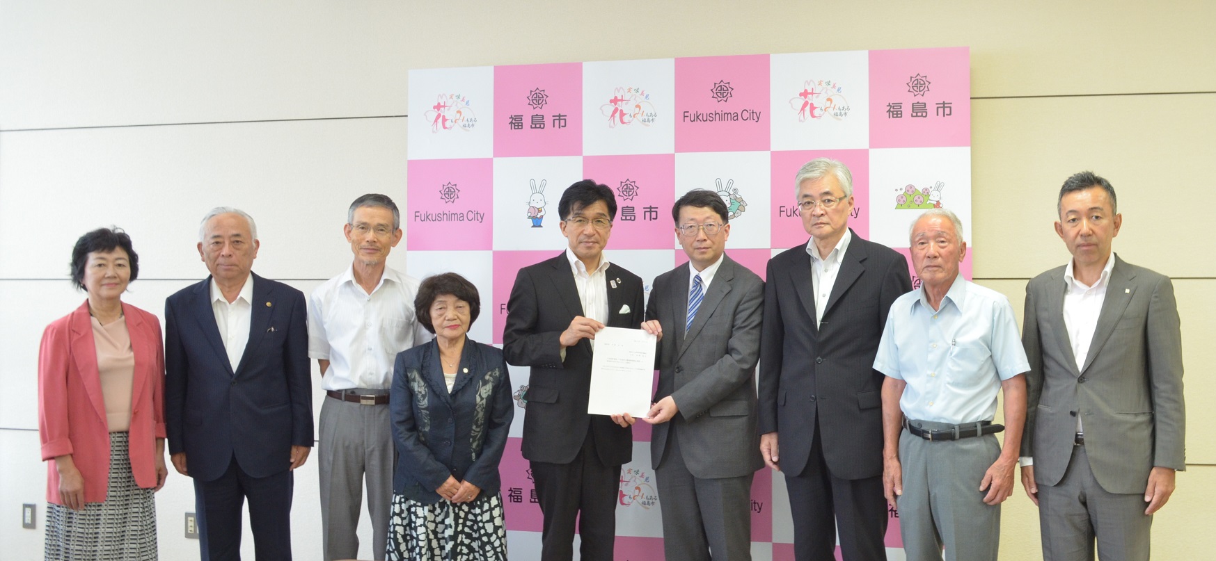 福島市のバックボードの前で、答申書を左右から持った市長とグレーのスーツの男性を中央にして男女7人の委員の方々が並んで写った記念写真