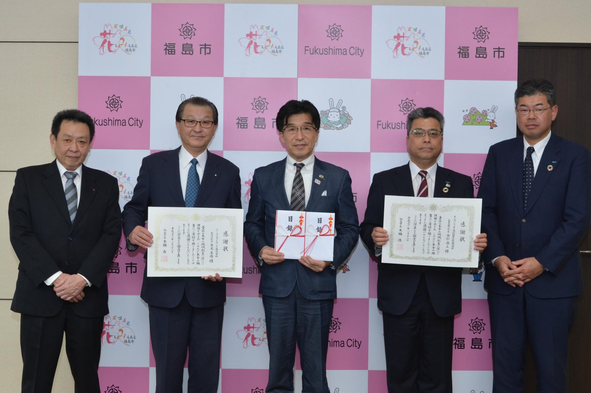 福島市のバックパネル前で、市長が目録を手に持ち、ジーエルサイエンス株式会社と、あいおいニッセイ同和損害保険株式会社の男性関係者が賞状を手に持っている写真