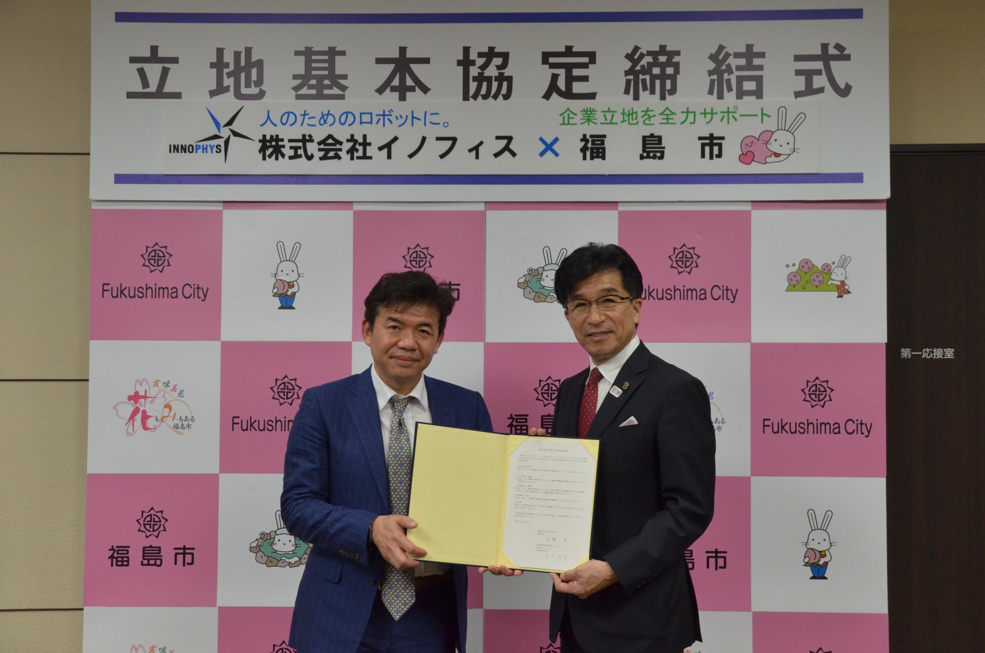 「立地基本協定締結式」と書かれたバックパネルの前で、一緒に協定書を持っている古川社長と市長の写真