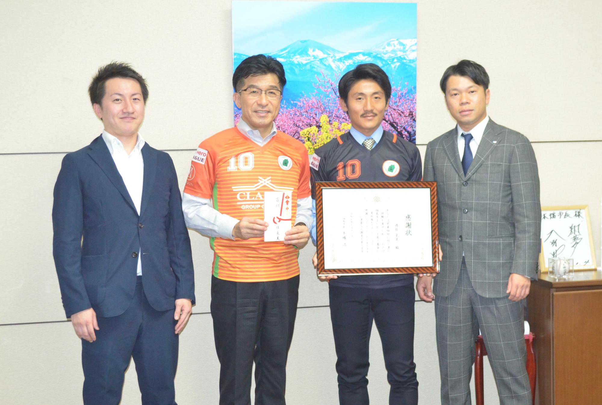 オレンジ色のサッカーのユニホームを着用し、金封を手に持った市長と3人の男性が横並びになり、1人が額縁に入った賞状を手に持ち記念撮影している写真