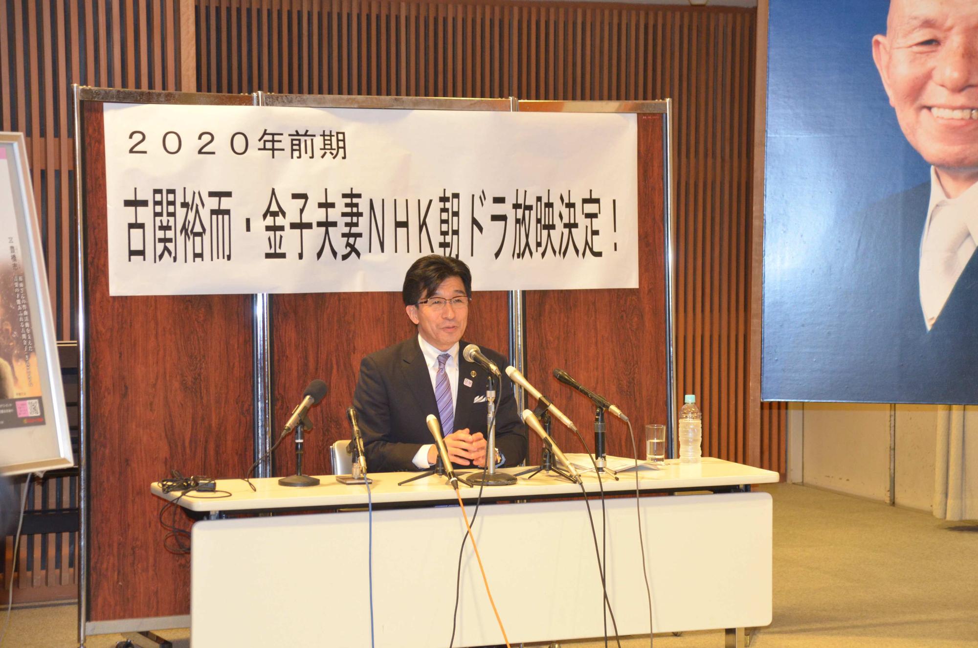 「古関裕而・金子夫妻NHK朝ドラ放映決定!」の横断幕の前で、椅子に座りマイクに向かって話をする市長の写真