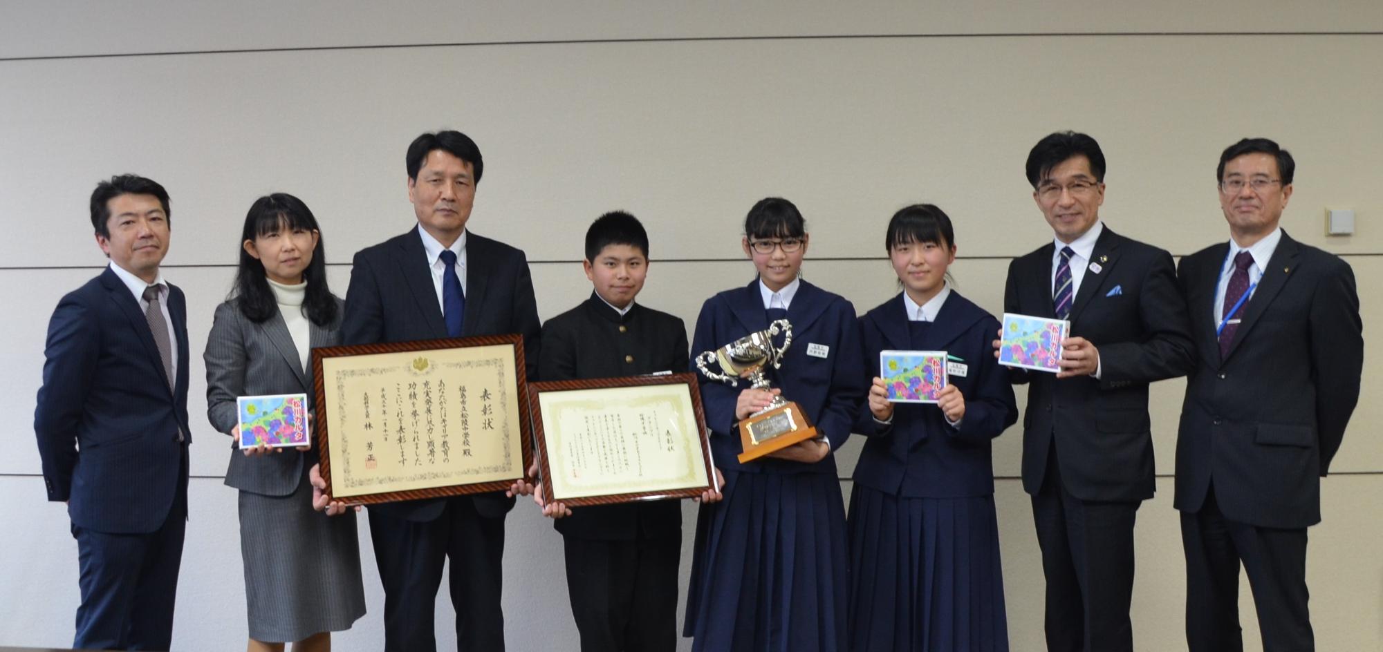 かるたや賞状、トロフィーを手に持った中学生と関係者7人と市長が、横一列に並んでいる写真