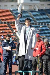 屋外の競技場で市長がライトグレーのコートを着て台に乗り、スターターピストルを掲げている写真