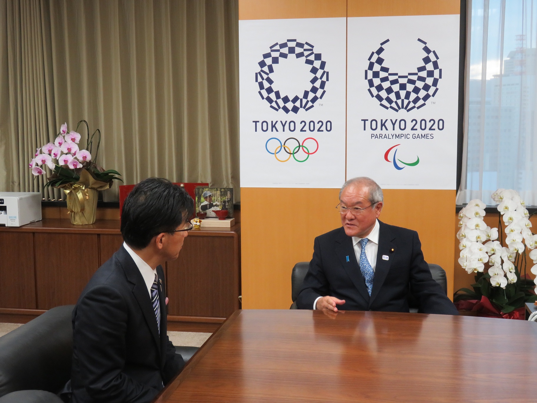 東京オリンピック、パラリンピックの組市松紋のマークが壁に飾られた部屋で、市長と男性が話している写真