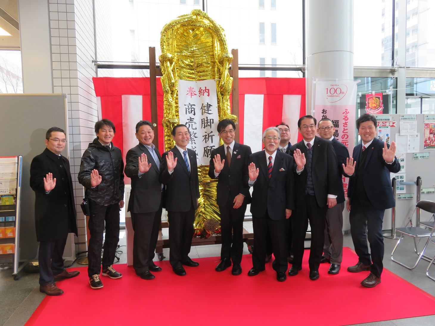 金色で作られ、奉納の幕がかけられた大わらじの前で、市長の他10名ほどの男性が広げた手のひらを前に向けて、ポーズをとっている写真