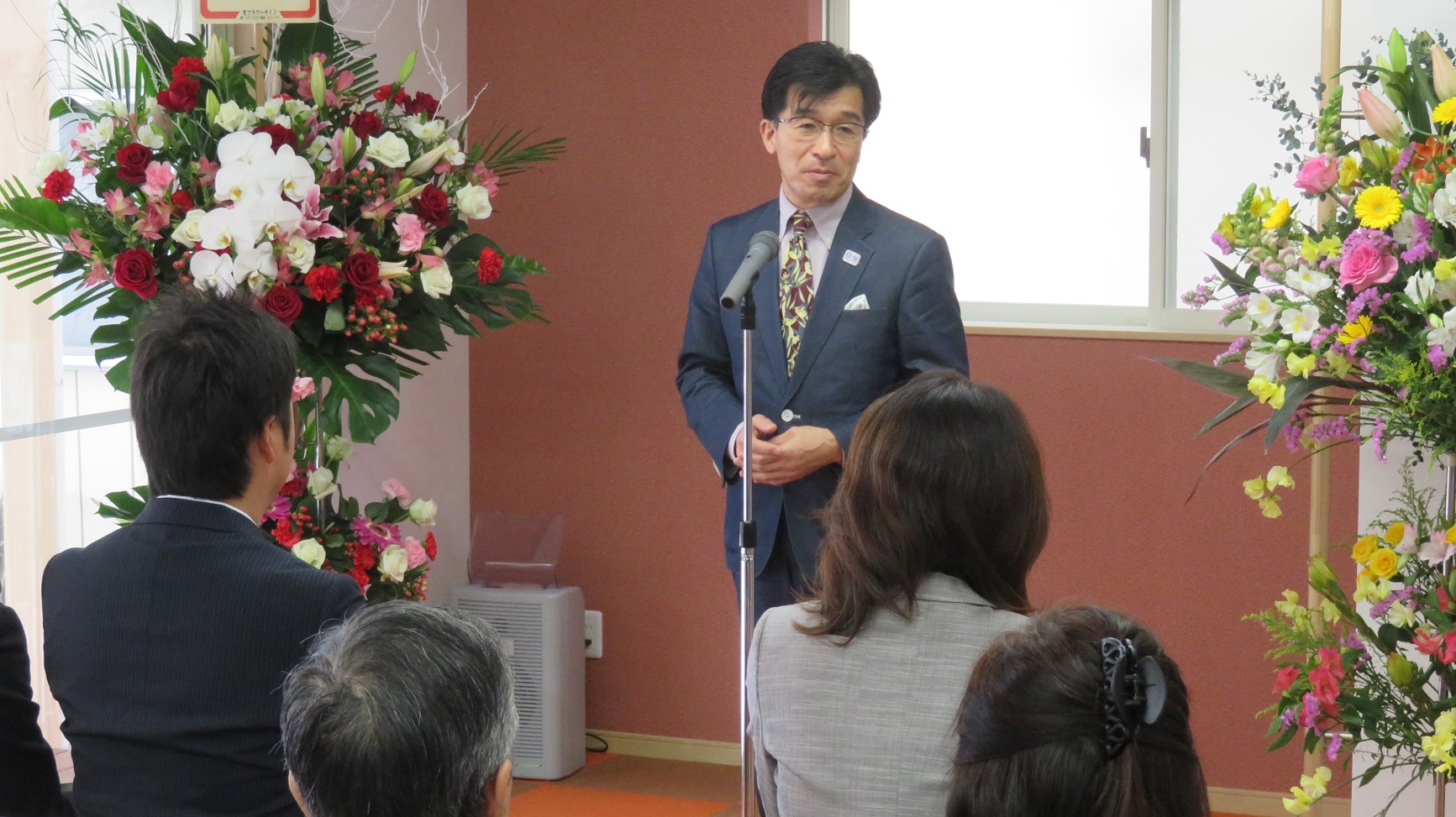 花が飾られた室内で、参加者たちの前に立ちスタンドマイクで話をしている市長の写真