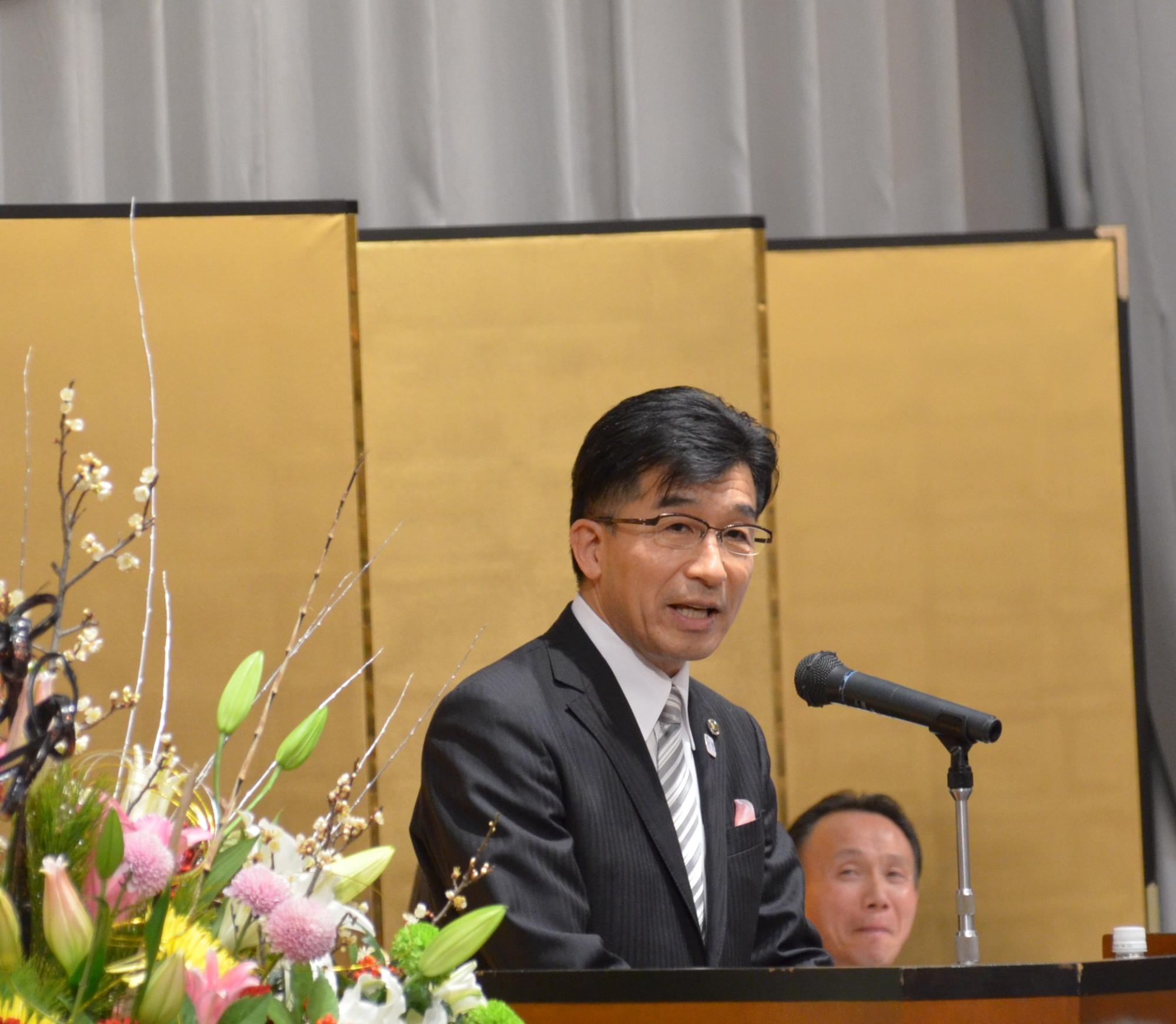金屛風の前の演壇でマイクスタンドの前に立ち、話をする市長の写真