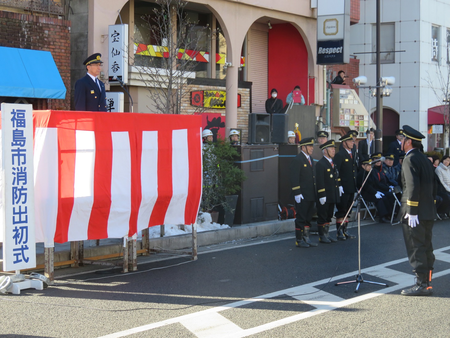 紅白幕の演説台に立つ男性と、消防団の制服を着た関係者がマイクスタンドの前に立ち整列している写真