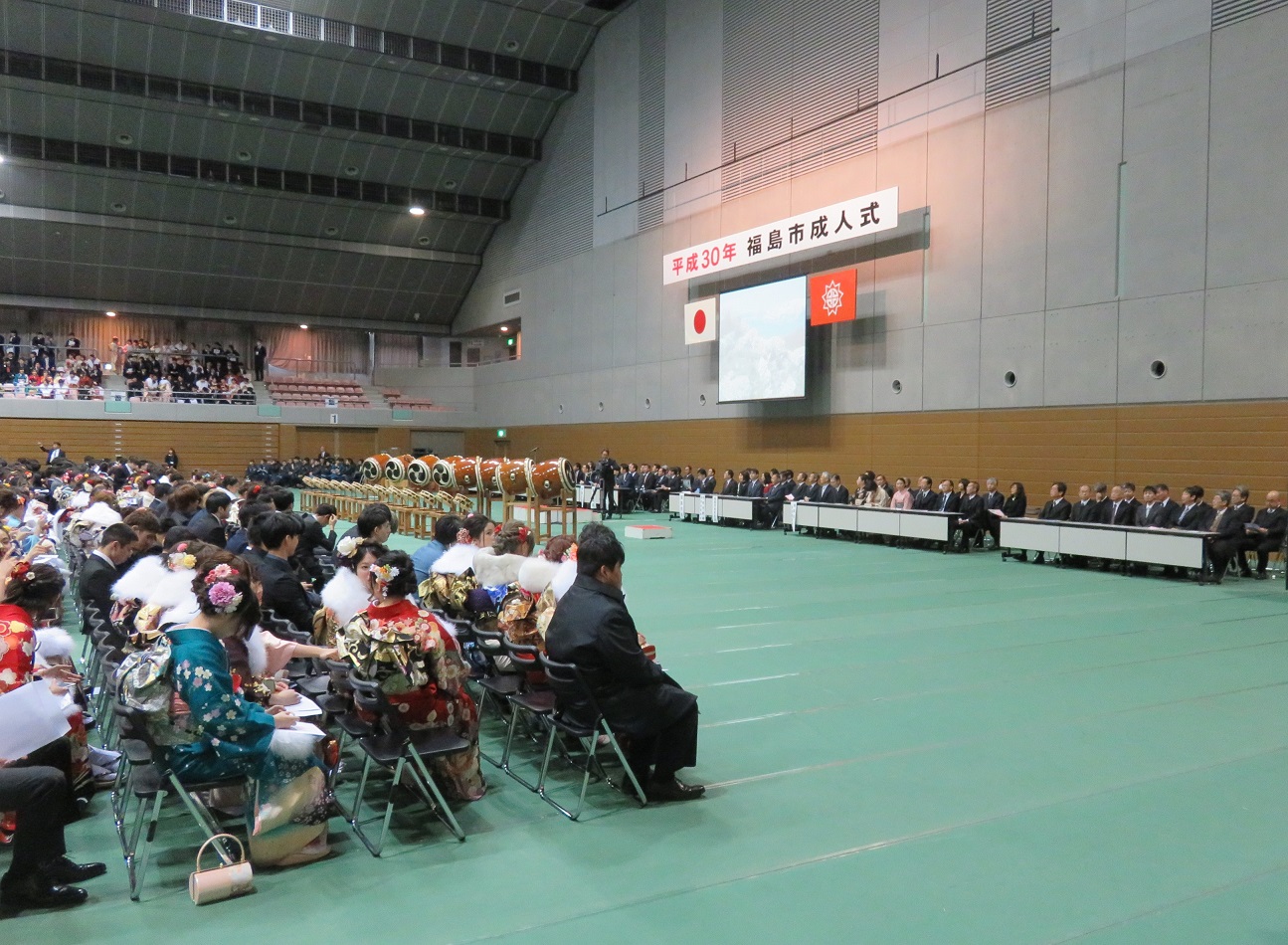 天井の高い広々とした体育館に「平成30年福島市成人式」の吊りパネルが掛けられており、来賓者と着物やスーツを着た男女が椅子に座っている写真